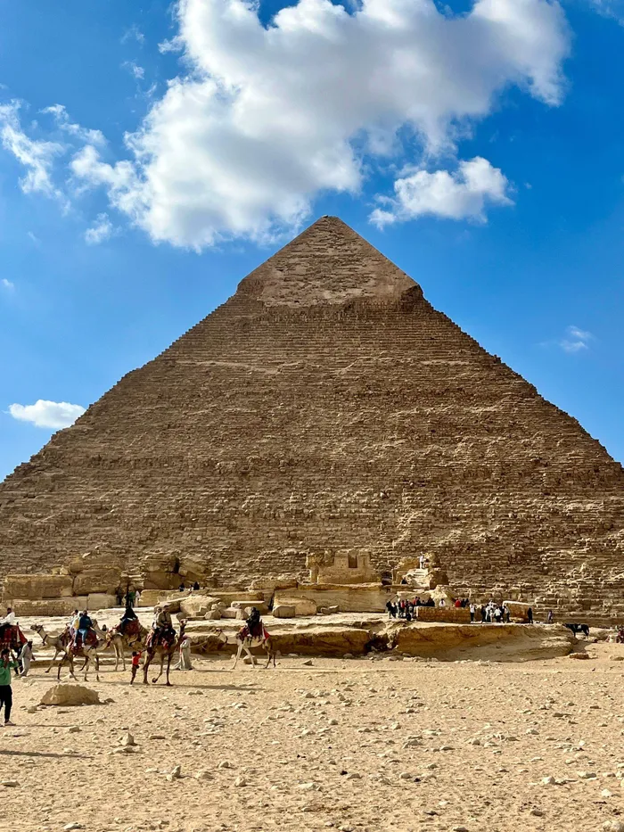 The legendary Great Pyramid of Giza stands prominently against a striking blue sky, accompanied by visitors and camels.

