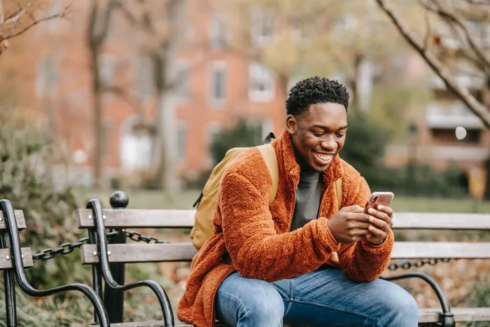 A young man sits on a bench smiling at his cellphone.