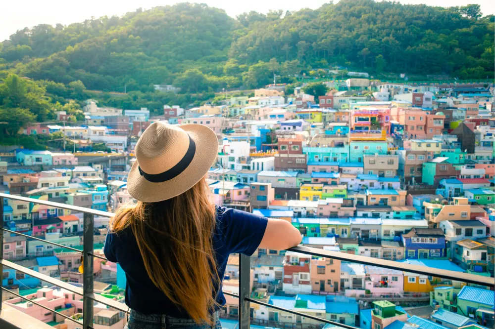 A young woman with long brown hair wearing jeans, T-shirt and a sunhat, leans on a railing overlooking a hillside of colourful small buildings.