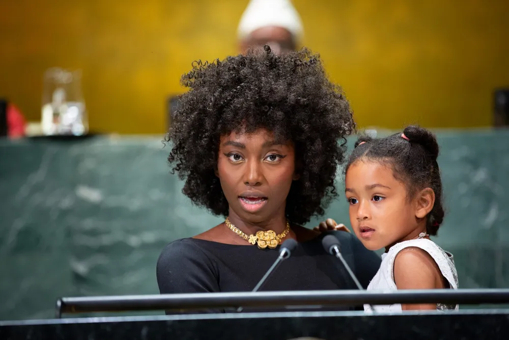 Woman with an afro and a baby on her arm wearing a gold necklace and black top. 