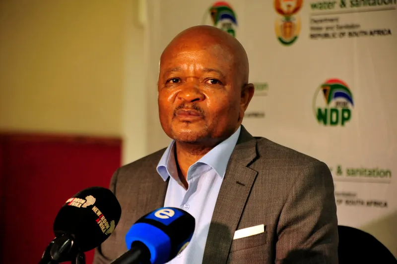 The minister sits before microphones with a department banner behind him.