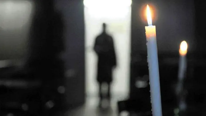 Two candles burn in a darkened room as a person is seen in the doorway.
