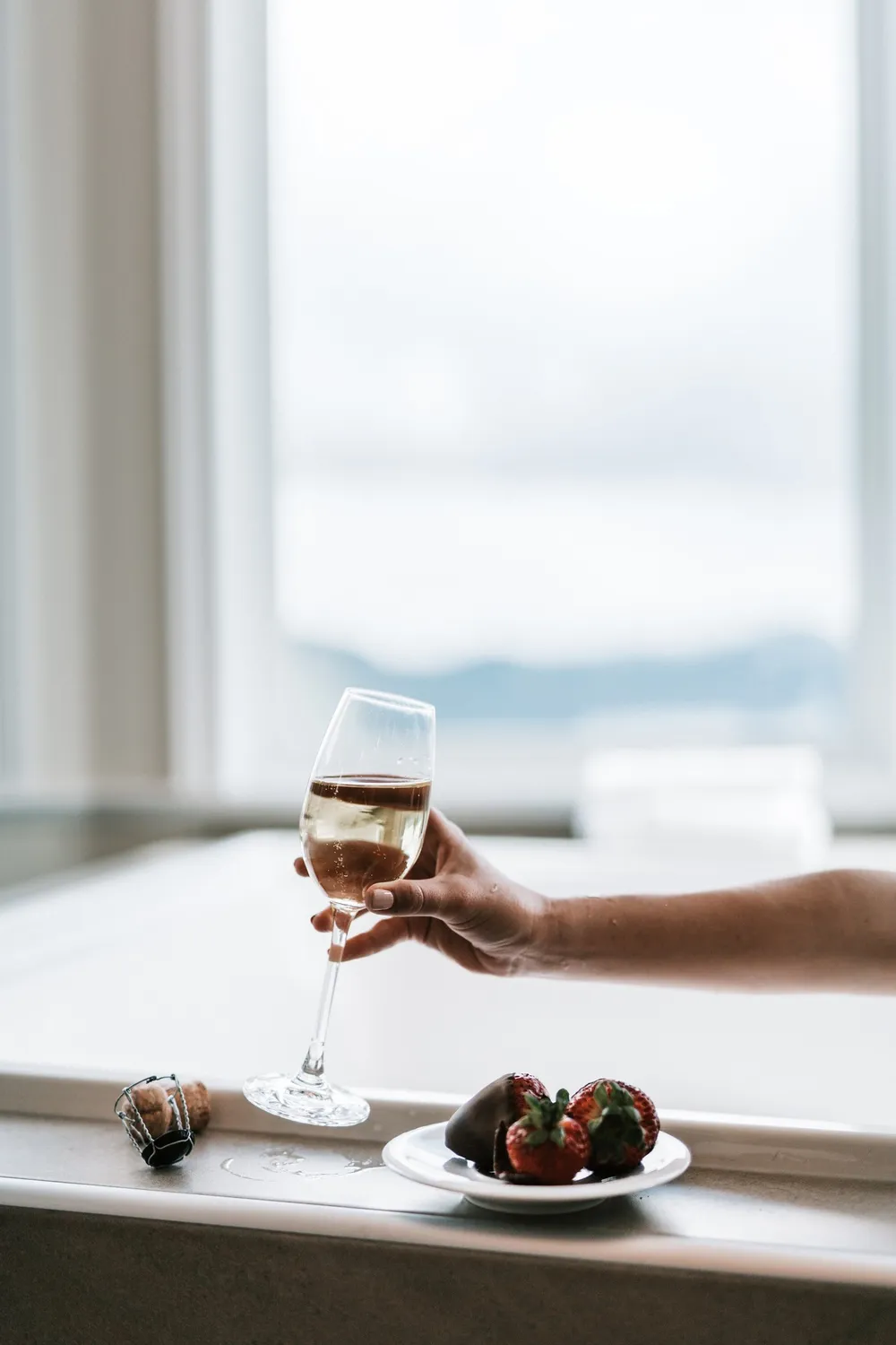 A hand lifts a glass of white wine from a ledge which holds a small plate of chocolate-dipped strawberries.