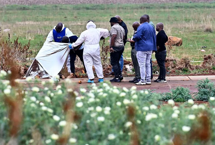 Police and a forensic investigator lift a silver sheet over the body of a woman found in Lama Street, Dawn Park, Boksburg, on Thursday, as detectives take notes beside them. She is the ninth woman found dead in Ekurhuleni in nine weeks.