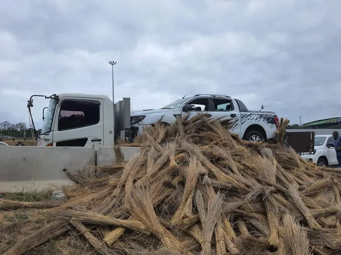 A white bakkie on the back of a truck behind a pile of traditional brooms.